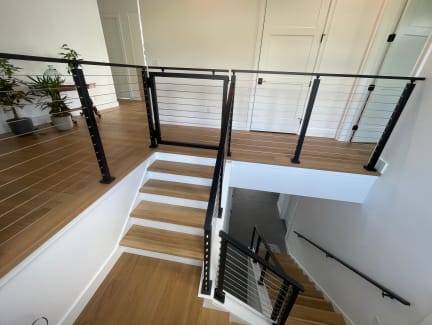 A black cable railing gate opens to oak stairs at a landing with a wall-mounted handrail below.