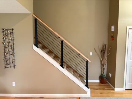 Black posts with cable infill and a wood cap rail atop a white knee wall beside a carpeted stair.