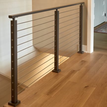 Dark bronze posts with cable infill and a matching top rail guard a stair opening on white oak floors.