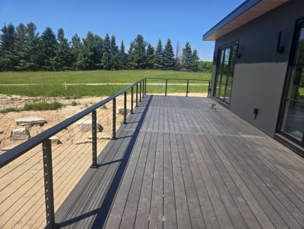 Black cable railing runs along a composite deck beside a dark modern home and evergreens