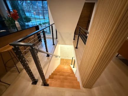 Black cable railing wraps a stair opening beside a wood-slat wall and kitchen island looking down a wood stair.