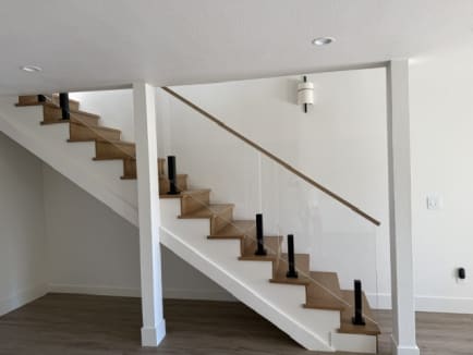 An oak staircase with glass panels on black ascend talons and a slim wood handrail in a white room.