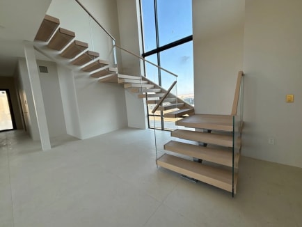 Light oak floating treads with frameless glass railing and wood handrail rise beside tall windows in a minimalist interior.