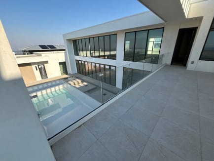 Frameless glass railing lines a stone-tiled upper terrace overlooking a pool courtyard in a modern white home.