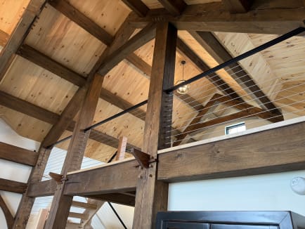 Cable railing with black top rail runs between heavy timber posts in a vaulted pine timber-frame home.