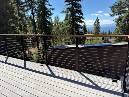 A black cable railing with wood cap edges a gray composite deck with pines and a distant lake view.