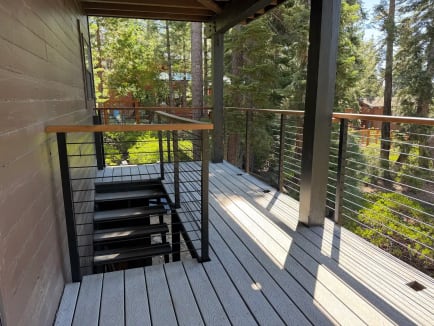 A covered gray deck with wood-capped cable railing and an open metal stair sits among pines.