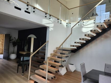Two black FLIGHT Mono stairs with thick oak treads rise to a glass balcony held by black standoff pins under a wood cap rail.