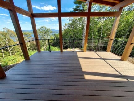 Black cable railing surrounds a composite deck under an angled timber-frame roof overlooking forested hills.
