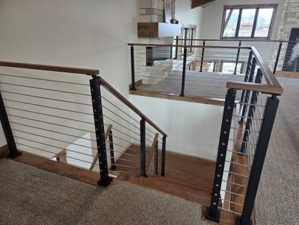 Black cable railing with wood caps wraps a stair landing overlooking a stone fireplace.