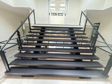 Dark wide floating treads on dual stringers with black-post glass railings rise toward a bright skylit hallway.