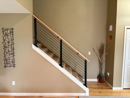 Black posts with cable infill and a wood cap rail atop a white knee wall beside a carpeted stair.