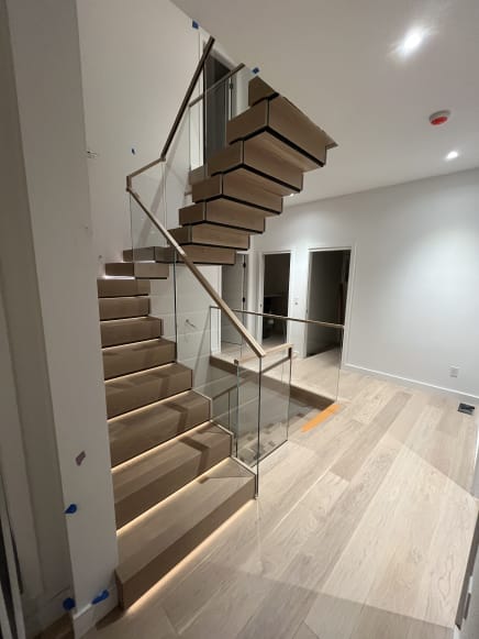 A U-shaped stack stair of thick white oak treads with LED step lighting rises between frameless glass guards with a wood handrail.