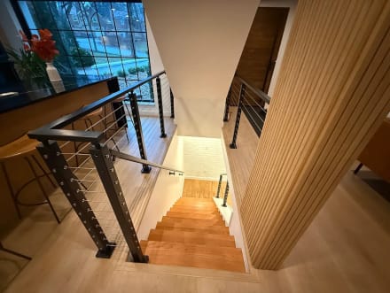 Black cable railing wraps a stair opening beside a wood-slat wall and kitchen island looking down a wood stair.