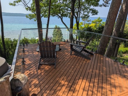 Frameless glass on spigots edges a wood deck among pines overlooking a bright blue lake.