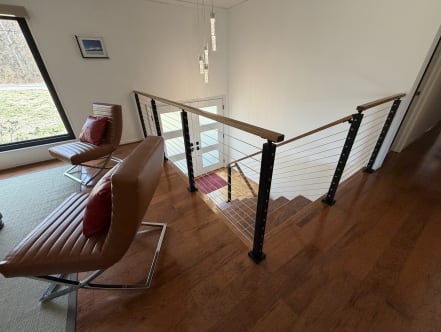 Black posts and a wood cap rail with stainless cables guard a stair opening beside leather lounge chairs and a large window.