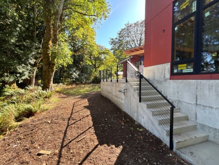 Black side-mounted posts with horizontal rods follow a concrete exterior stair beside a modern red-clad home.