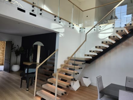 Two black FLIGHT Mono stairs with thick oak treads rise to a glass balcony held by black standoff pins under a wood cap rail.