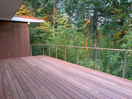 Stainless cable railing with a wood top rail edges a new hardwood deck surrounded by tall pines.