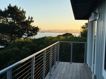 Silver-post cable railing on a wood balcony overlooking trees and water at sunset.