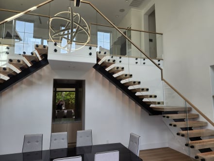 Two mirrored mono stringer stairs with standoff-pin glass railings meet a central landing above a dining room.