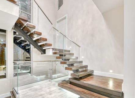 Wood floating treads on a dark mono stringer descend to a wide landing tread beside frameless glass with a wood handrail.