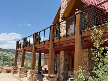 Black cable railing tops a timber-post deck on a stone-and-log mountain home under a blue sky.