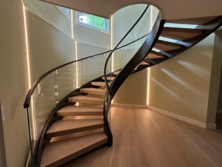 Sweeping curved stair with wood treads and dark stringers, glass railing and a curved dark handrail, lit by vertical wall LED strips.