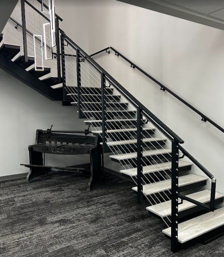 Black mono stringer switchback stair with gray wood-look treads, black cable railing and wall handrail in an office lobby.