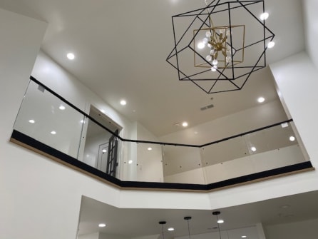 Glass railing with black base and top cap lines an open upper-floor balcony below a geometric chandelier.