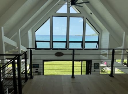 Black horizontal rod railing along an open loft under a white vaulted ceiling with a triangular wall of windows facing the ocean.