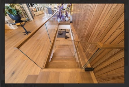 Frameless glass panels with a wood handrail line a wood-clad stairwell descending from a white oak floor.