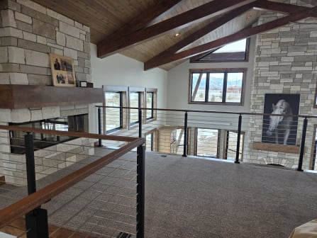 Black cable railing with a wood cap overlooks a vaulted great room with a stone fireplace and timber beams.