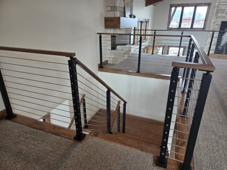 Black cable railing with wood caps wraps a stair landing overlooking a stone fireplace.