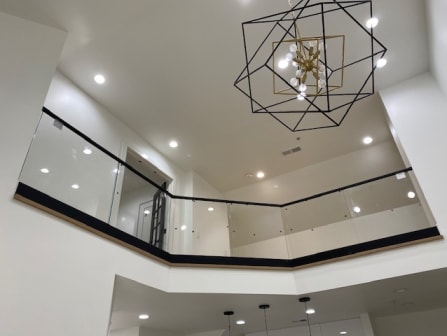 Glass railing with black base and top cap lines an open upper-floor balcony below a geometric chandelier.