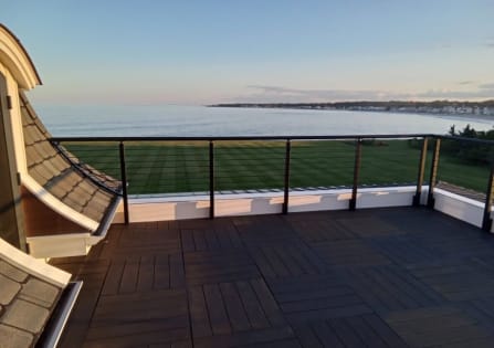 Black cable railing with a curved section borders a dark rooftop deck overlooking a lawn and the ocean at dusk.