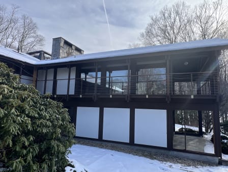 A dark-stained timber two-story home with a long upper balcony enclosed by horizontal stainless cable railing, snow on the ground.