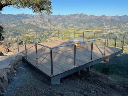 Brushed stainless cable railing around a wood deck perched above a vineyard valley under blue sky.