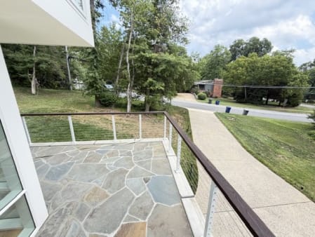 Cable railing with white posts and wood rail wraps a flagstone terrace overlooking a tree-lined street