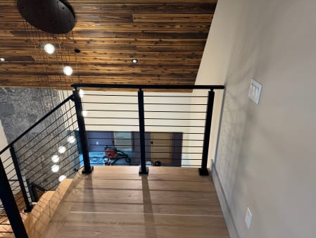 Black cable railing wraps a stair opening beneath a reclaimed-wood ceiling and globe pendants