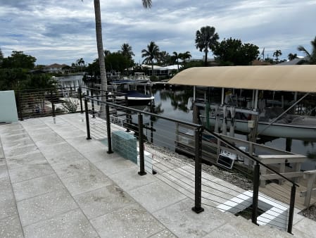 Black cable railing runs along a paver patio beside a canal with boat docks and palms.