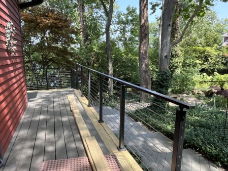 A gray deck beside a red house edged with black cable railing amid lush trees.