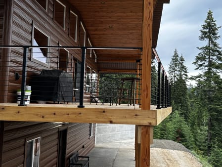 Black cable railing wraps an elevated cedar deck on a log-sided cabin surrounded by tall pines.