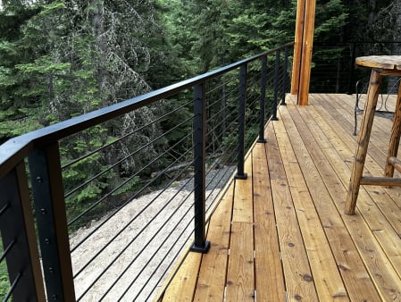 Black cable railing with flat top rail lines a cedar deck beside a rustic table in a forest setting.