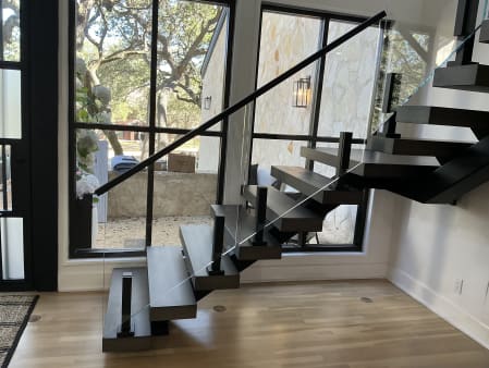 Dark-stained floating treads turn at a landing on a black mono stringer with frameless glass on talons before tall windows.