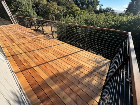 Black posts with wood cap and stainless cables wrap the corner of a new wood deck above green brush.