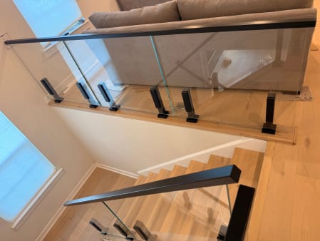 Glass panels on black talon clamps with a black handrail guard a stair opening and landing.