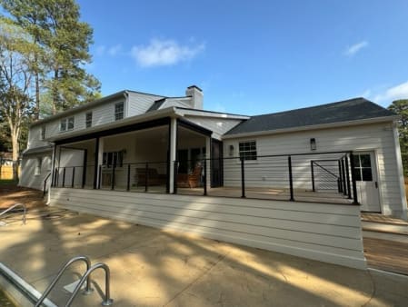 Long raised porch with black rod railing overlooks a pool deck behind a white home.