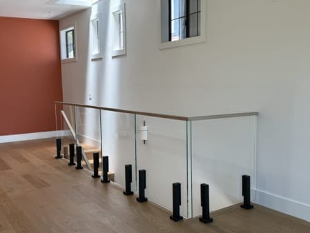 Black talons hold frameless glass with a white oak cap around a stair opening in a bright upstairs hall.