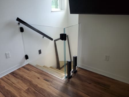A frameless glass gate with black hinges guards the top of a stair beside a black wall handrail.